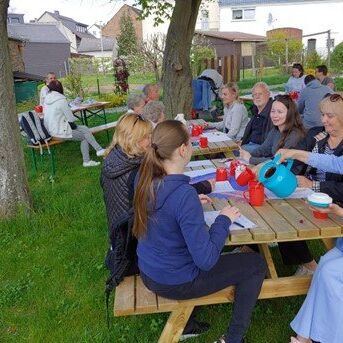 Männer und Frauen sitzen an Holztisch im Pfarrgarten mit Getränken
