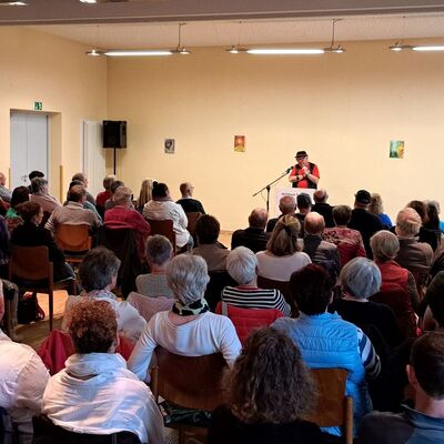 Mann auf Bühne mit Mikrofon, davor sitzen viele Menschen auf Stühlen