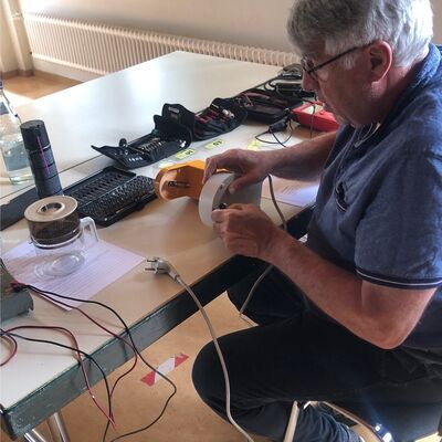 Älterer Mann mit Brille sitzt mit diversen Werkzeuguntensilien an einem Tisch und repariert eine Filterkaffeemaschine