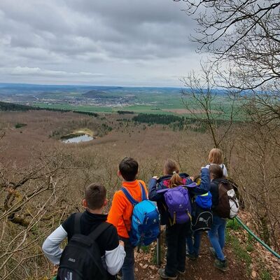KInder mit Rucksächen stehen an einem Aussichtpunkt mit Geländer und überblicken Wald ohne Laub, einen See und weite Landschaft und Ort am Horizont