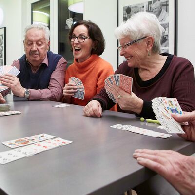 Am Tisch sitzend ein älterer Mann und zwei lachende Frauen halten Spielkarten in der Hand, ein Händepaar im Anschnitt hält ein Kartenblatt in der Hand