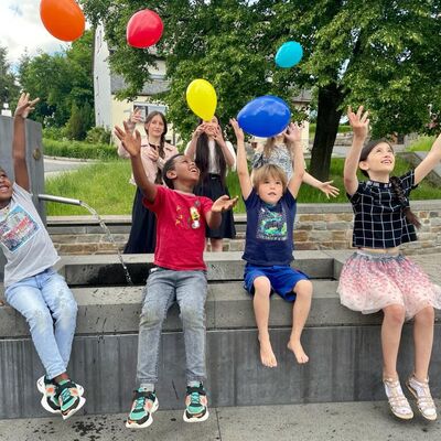 Kinder siten lachend auf einer Bank im Freien und spielenm mit bunten Luftballons