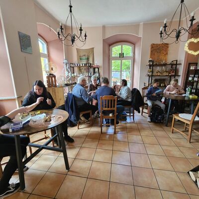 Bild mit Blick in einen Ladenraum, in dem sich mehrere Tische befinden an denen Personen unterschiedlichebn Alters verschiedene Spiele spielen, im Vordergrund ein Holzofen