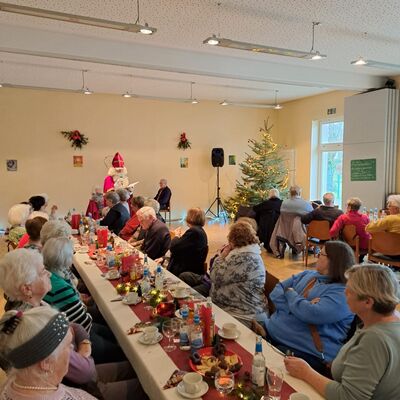 Senior*innen sitzen an langen weihnachtlich geschmückten Tafeln mit Kerzen und hören einem Nikolaus zu, daneben steht ein Weihnachtsbaum