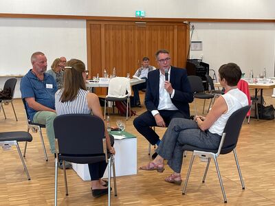 Minister spricht ins Mikrofon, 1 Mann und 2 Frauen sitzen rund um ihn, im Hintergrund hören weitere Personen zu