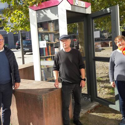 Bild mit zwei Männern und einer Frau, die vor einer Telefonzelle stehen, in der sich Regale mit Büchern befinden.