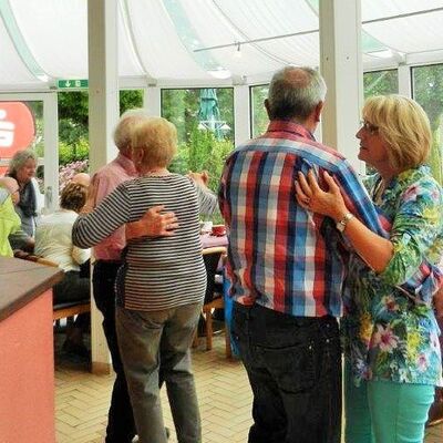 zwei tanzende Paare in einem Wintergarten, im Hintergrund sitzen Menschen an einer Bar und an Tischen und unterhalten sich
