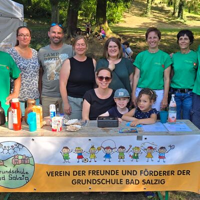 Gruppenbild mit Männer und Frauen mittleren Alters sowie Kindern hinter einem Tisch mit Thermoskannen, Kuchen und Tischkasse