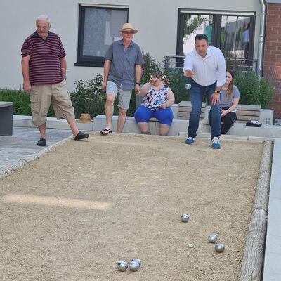 Menschen in Sommerkleidung: Drei Männer stehen an einem Boule-Feld mit Sand, darauf liegen Metall-Kugeln, ein Mann wirft eine Kugel. Im Hintergrund sitzen zwei Frauen auf einer Bank