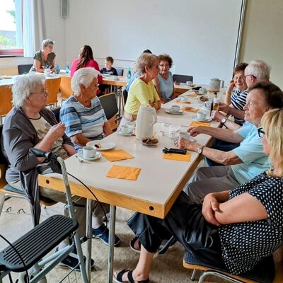 Bild mit älteren Menschen, einem Kind und zwei jüngeren Frauen, die in einem Gemeinschaftsraulm bei geckektem Tisch mit Kaffee und Kuchen sitzen, im Vordergrund ein Rollator.