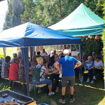 auf zwei Grills liegen Steaks und Würstchen, dahinter sind Zelte aufgebaut in denen Ältere in Sommerkleidung sitzen und stehen