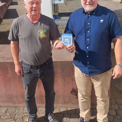 Zwei Männer halten gemeinsam ein Schild mit einem QR-Code in die Kamera