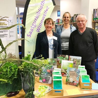 Bild mit zwei Männern und einer Frau die in einer Bibliothek hinter einem Tisch steehen, auf dem Infomaterial swowie Samentütchen und Pflanzen stehen, im Hintergrund ein Banner mit der Aufschrift Saatgutbibliothek.