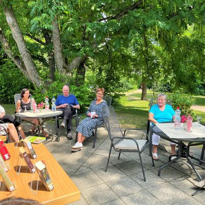 Auf einem Tisch stehen mehrere Bücher in Aufstellern, gegenüber sitzen Menschen auf der Terasse in der Sonne