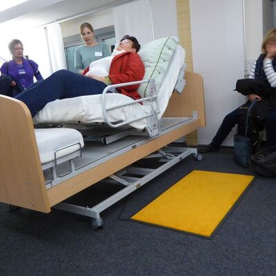 Bild einer Gruppe von Menschen um ein Pflegebett. Frauen und Männer stehen und sitzen um ein barrierefreies Bett, in dem eine ältere Frau liegt. Eine Frau erklärt dazu etwas.