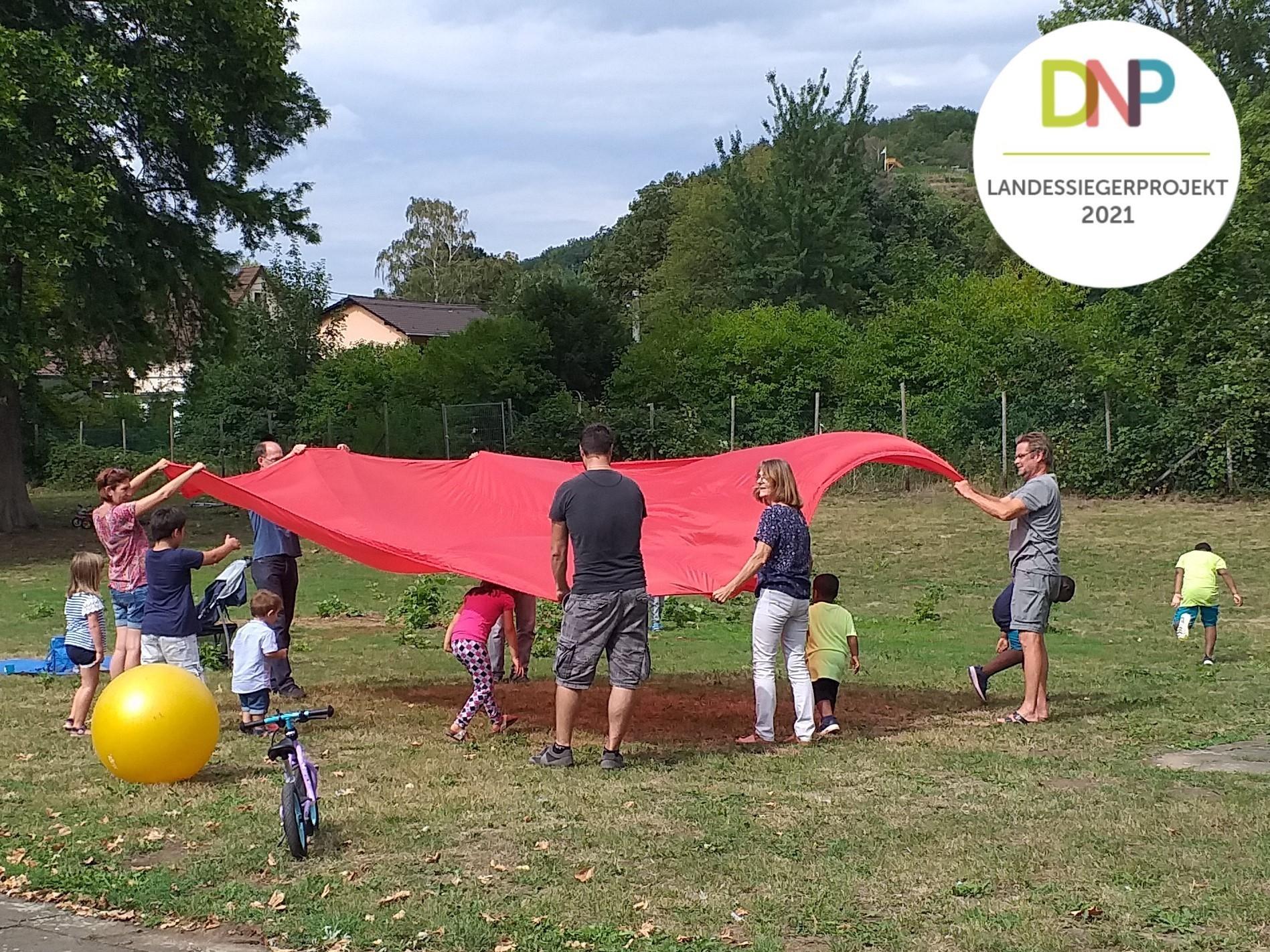 Gruppe von 6 Erwachsenen im Park spannt ein großes rotes Tuch auf, darunter laufen Kinder hindurch