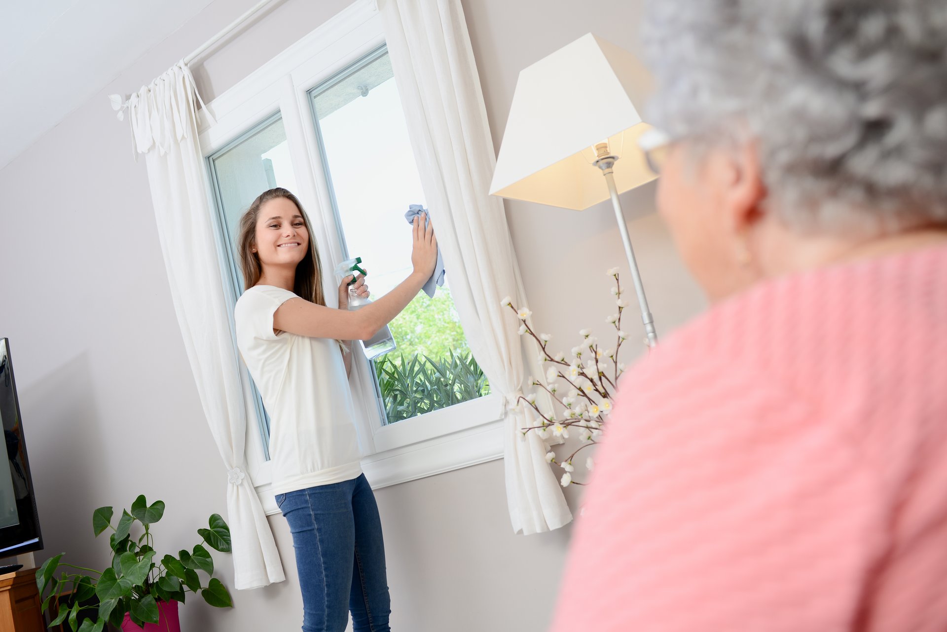 Junge Frau steht lachend an einem Fenster, das mit einen Lappen putzt, Alte Dame schaut ihr dabei zu.