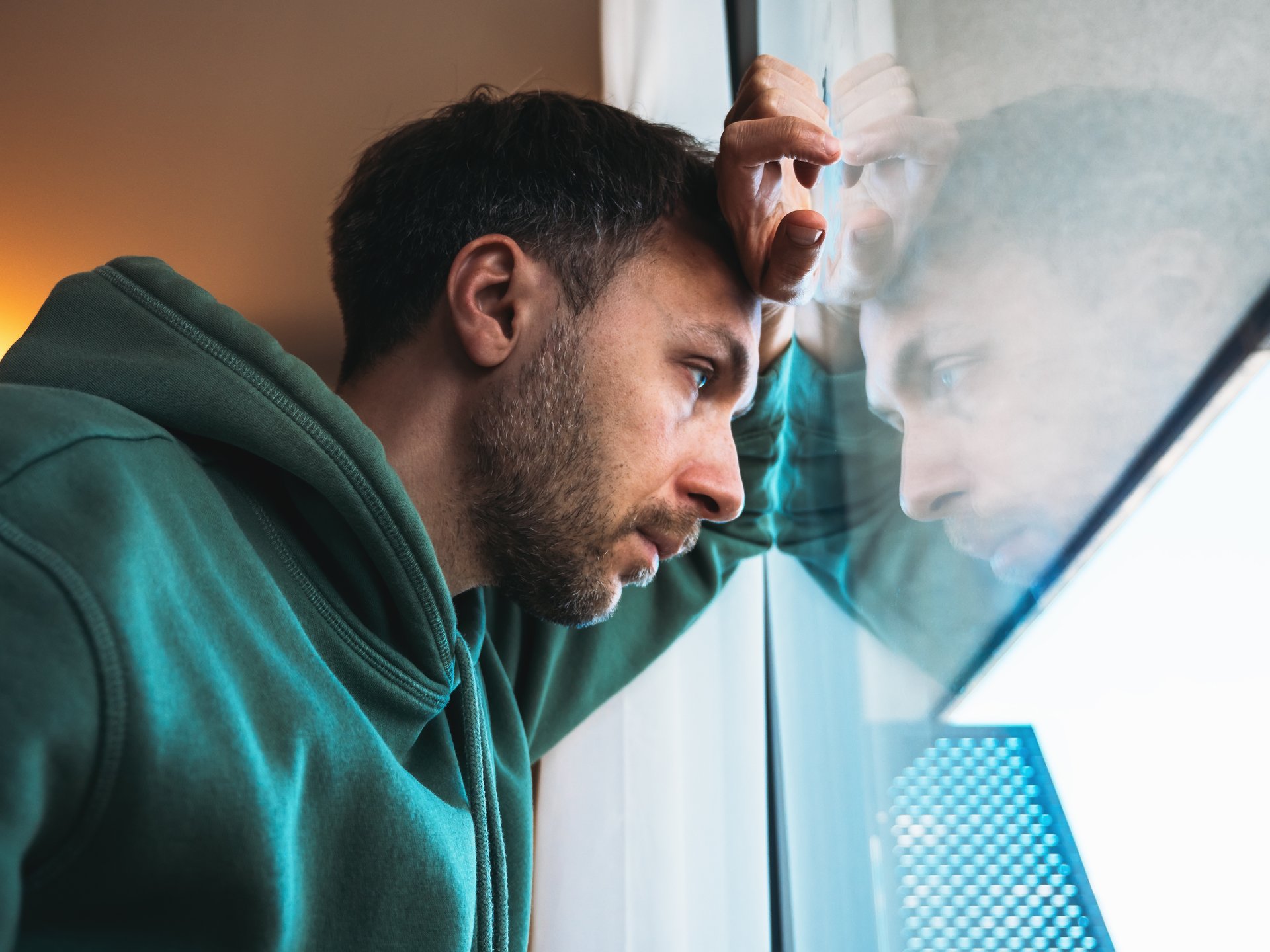 Bild mti einem mittelalten Mann, der traurig schaut und der mit Hand und Kopf an einem Fenster lehnt