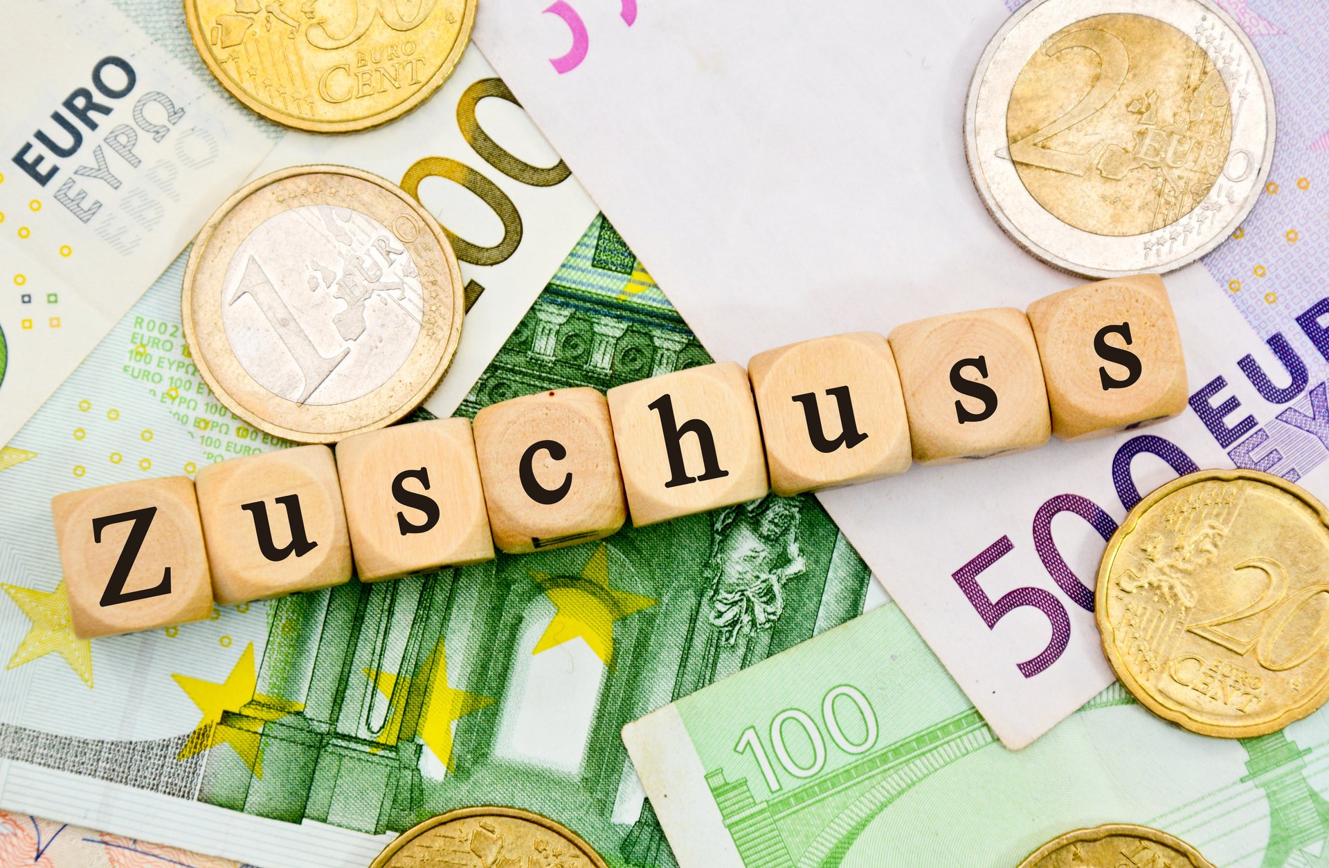 Aufgrünem 100 Euroscheinen und violetten 500-Euroscheinen liegen Euromünzen und Holzklötzchen mit je einem Buchstaben aneinandergereiht, die das Wort Zuschuss ergeben d