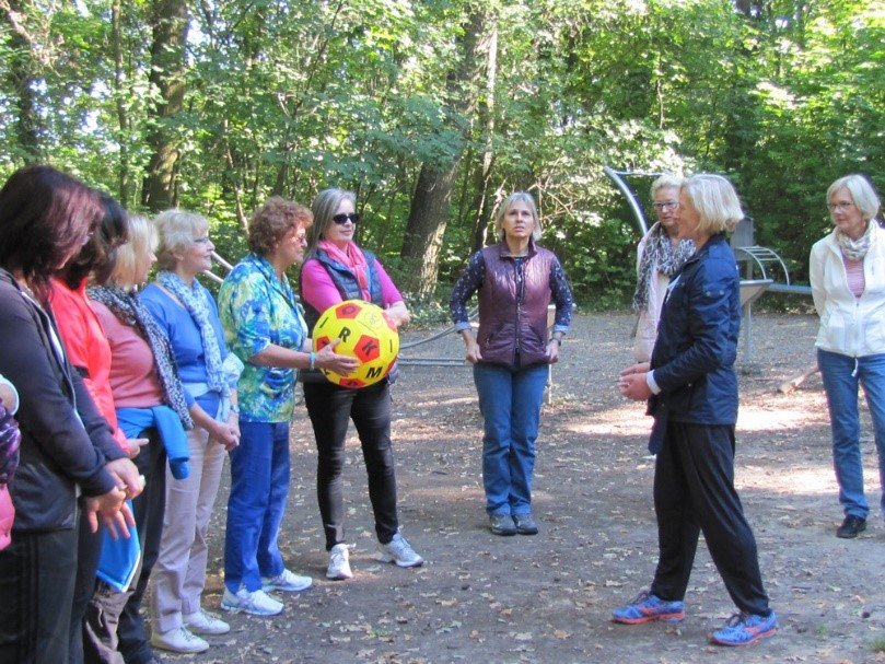 Gruppe von älteren Frauen im Park. Eine Trainerin erklärt mithilfe eines Balls auf dem Buchstaben abgebildet sind, den Teilnehmerinnen eine mögliche Bewegungsübung für ältere Menschen.